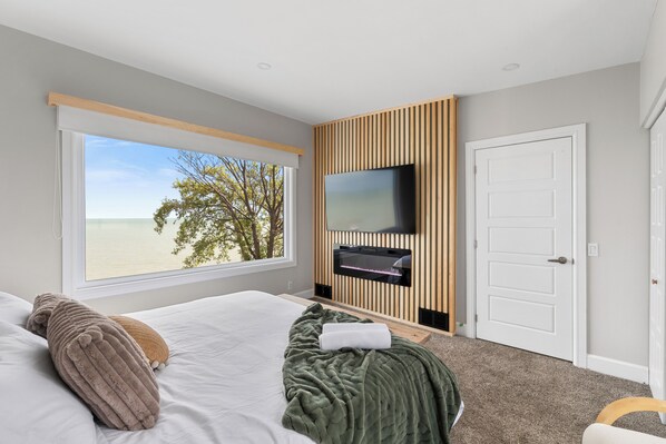 Un dormitorio con un ventanal grande, un televisor de pantalla plana, una cama con almohadas y una puerta.