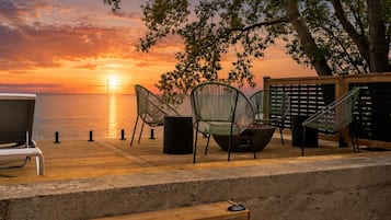 A wooden deck with outdoor seating, a fire pit, and a view of the sunset over the water.