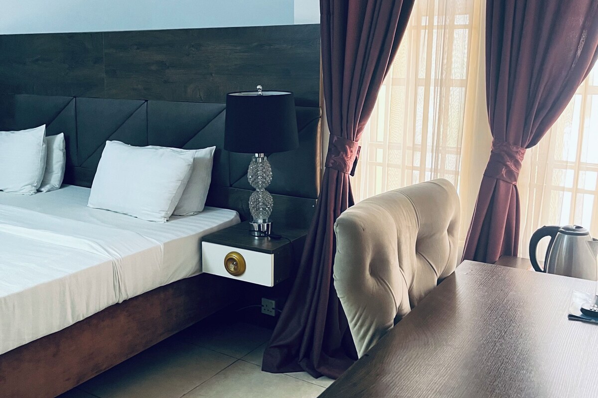 A hotel room with a bed, bedside table, lamp, and a view through curtains.