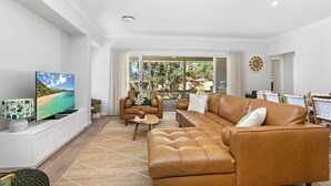 A modern living room with a large leather sofa, a flat-screen TV, and a dining area with wicker chairs. - Poppy's - Fire Pit - 4 Mins Walk to Beach (Callala Beach)