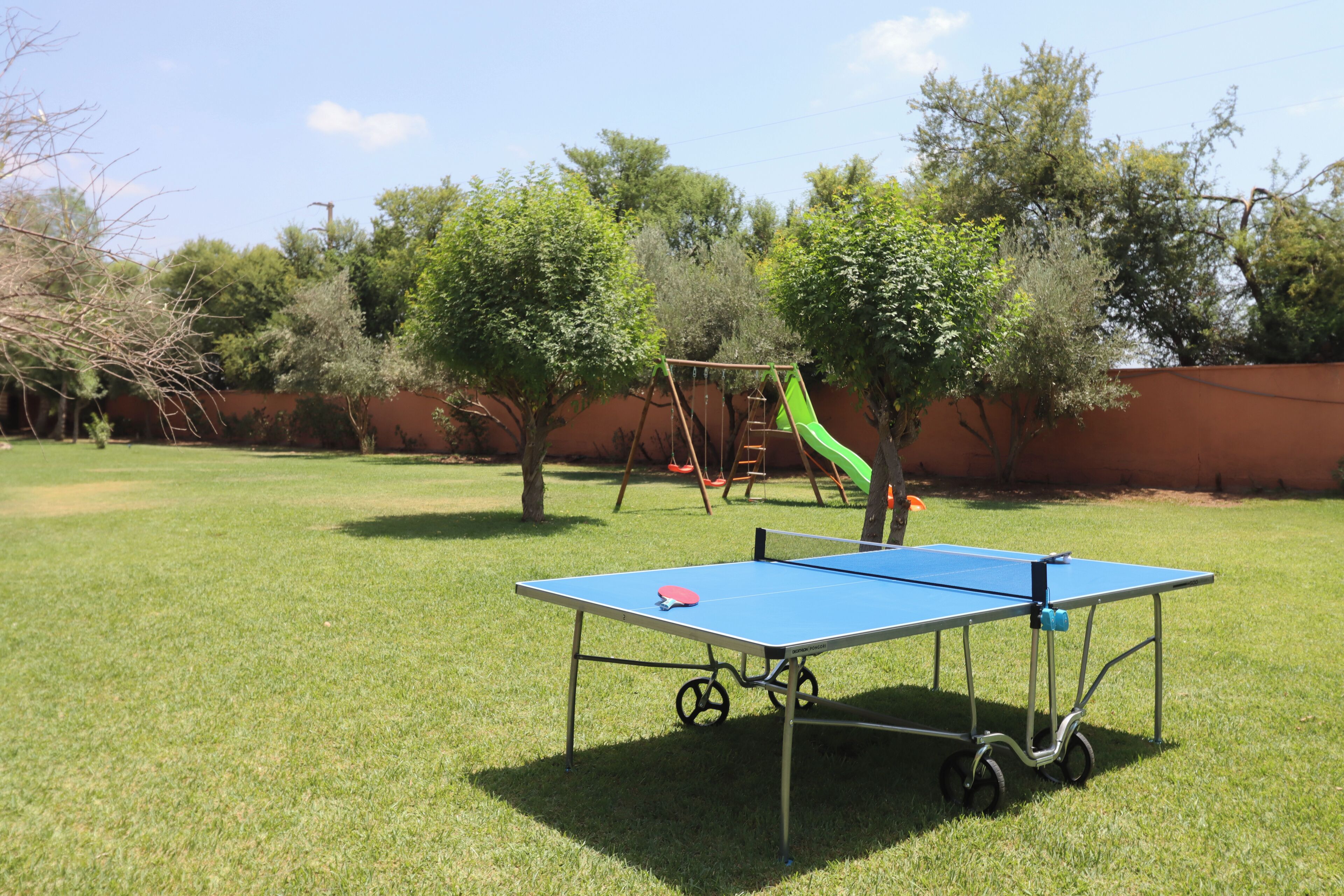 A blue table tennis table with wheels, a swing set, and trees in a grassy yard.