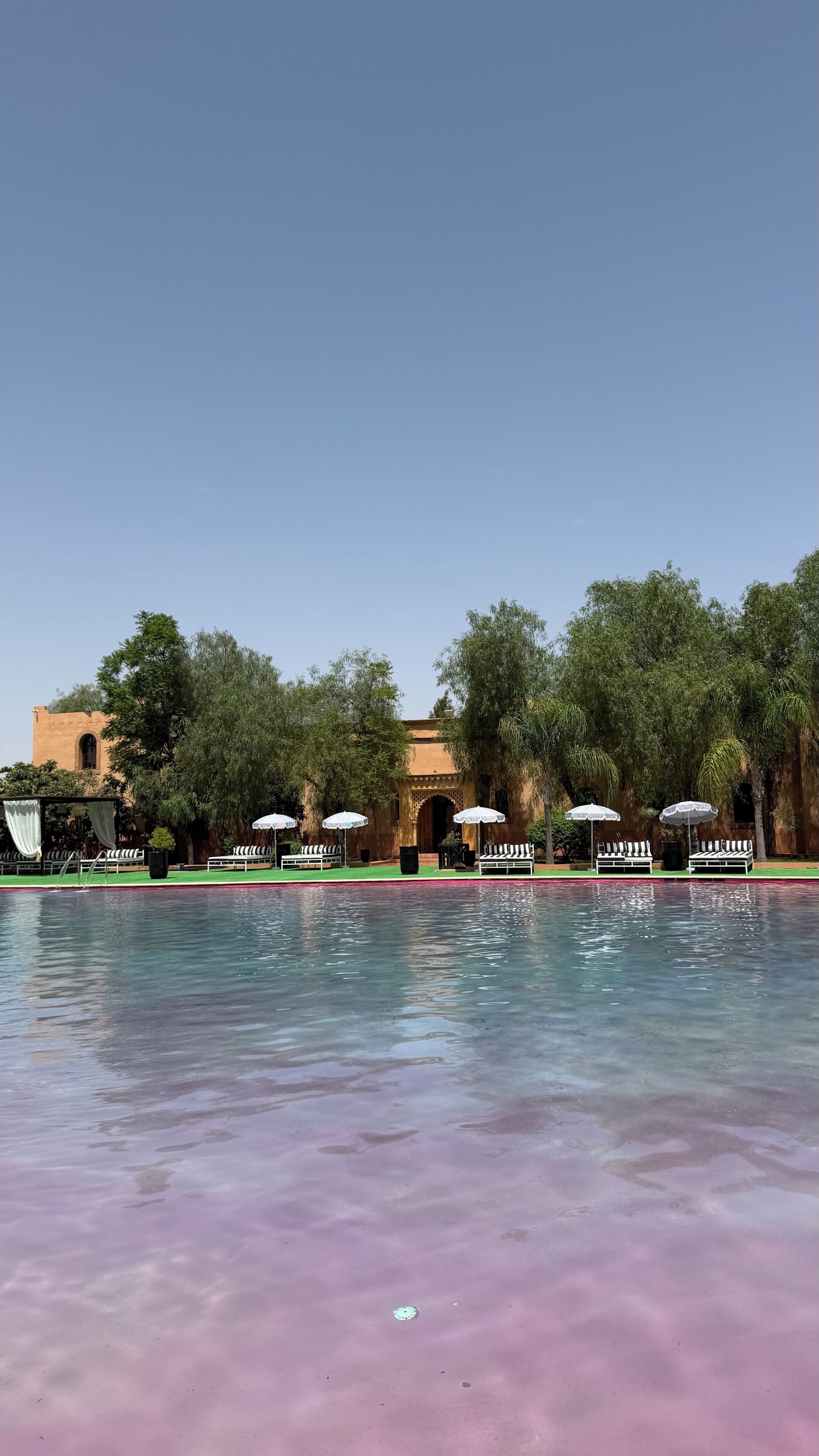 Zona de piscina con sillas de descanso y sombrillas, rodeada de árboles y un edificio con arcos.