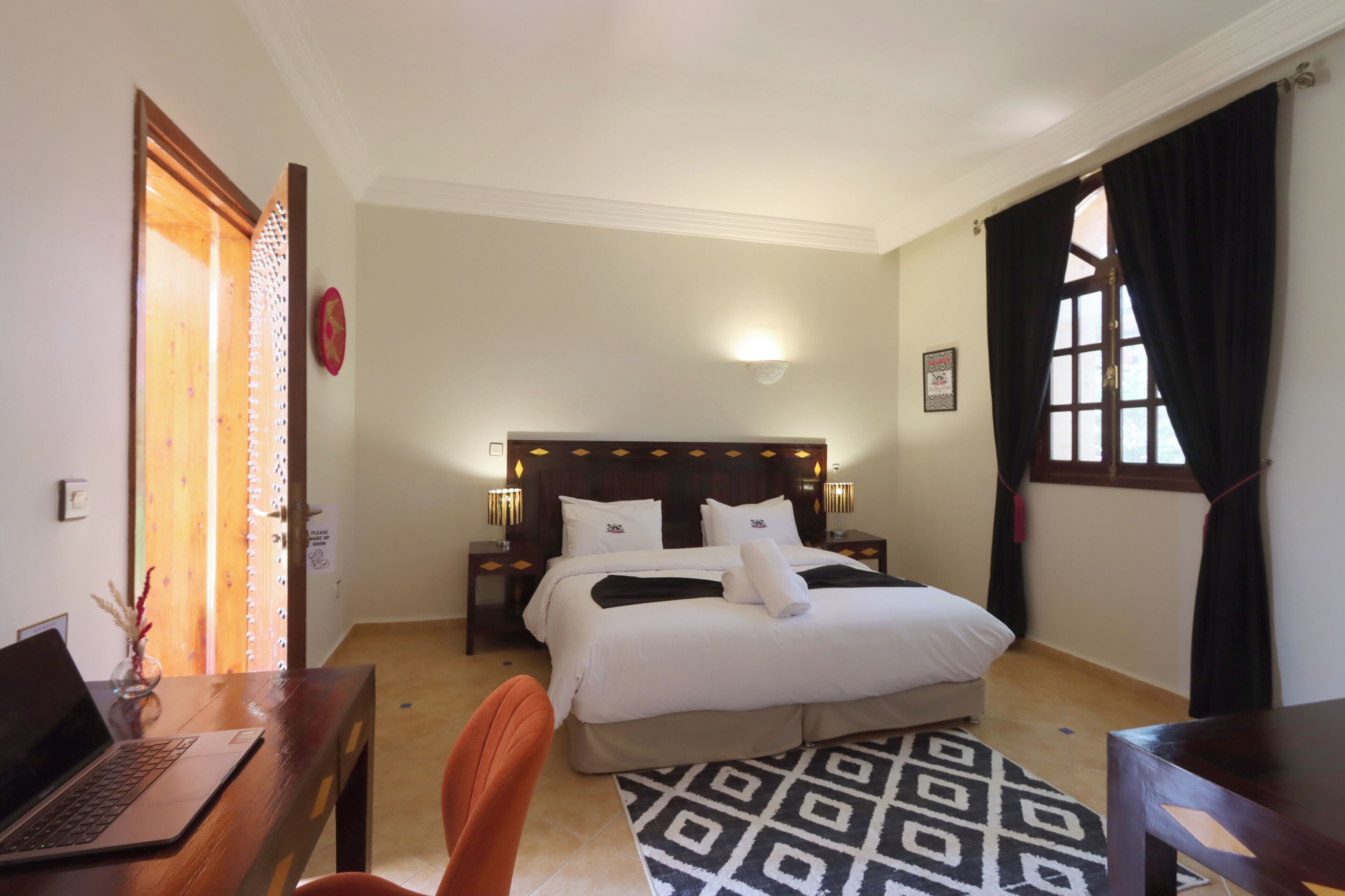 Ein Hotelzimmer mit Bett, Schreibtisch mit Laptop, Stuhl, Nachttisch, Lampe, Fenster mit Vorhängen und einer Wandleuchte.