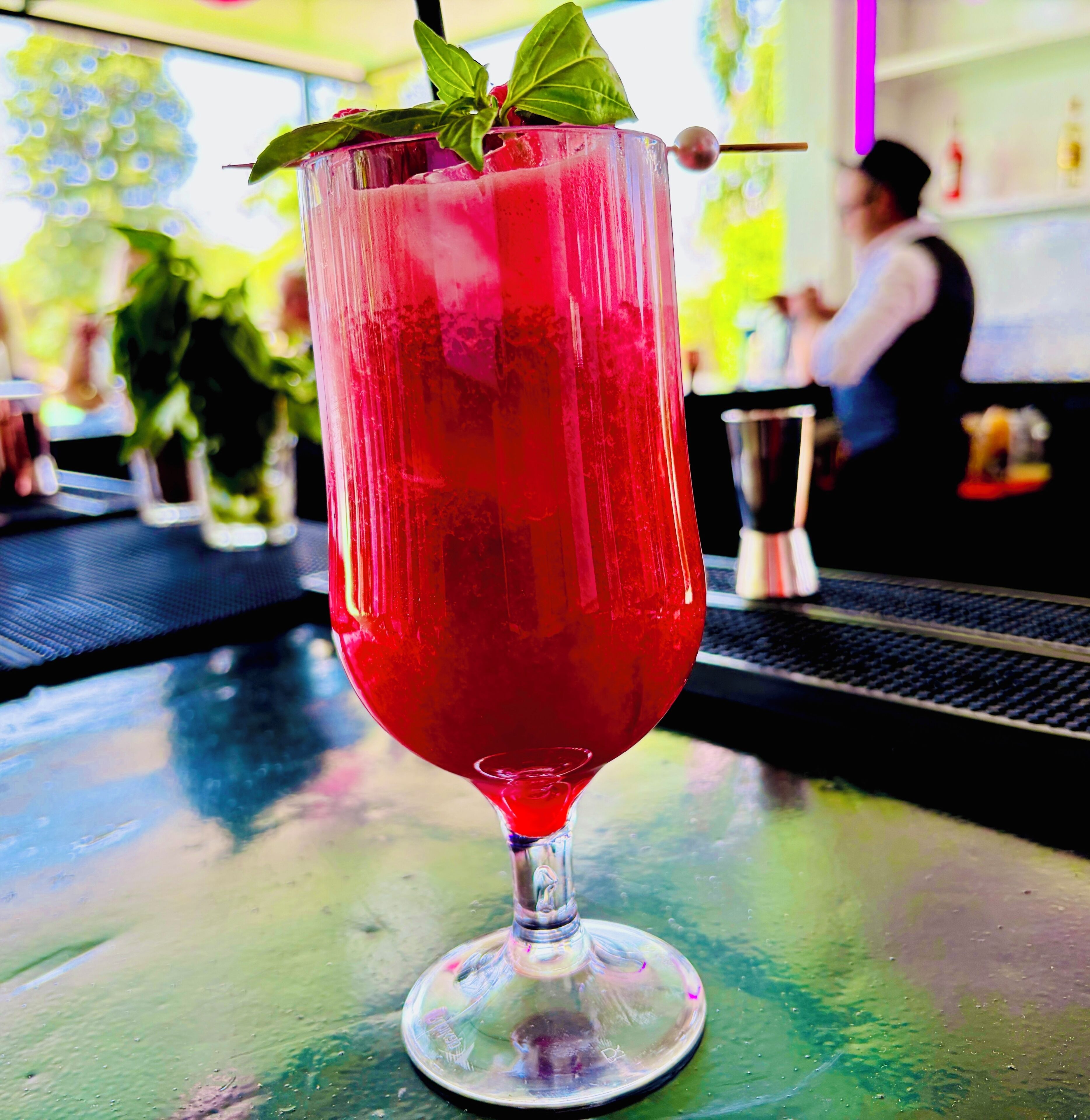 Un vaso alto con una bebida roja, adornado con una hoja, sobre una barra.