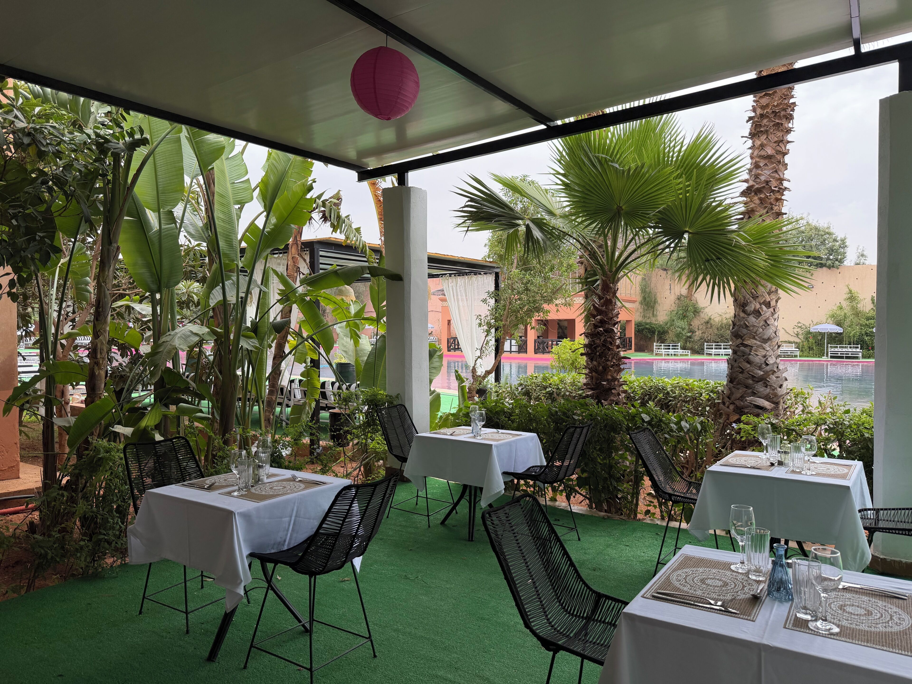 Un área de comedor al aire libre con mesas y sillas, rodeada de abundante vegetación y vista a una piscina.