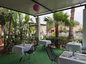 An outdoor dining area with tables and chairs, surrounded by lush greenery and a view of a swimming pool.