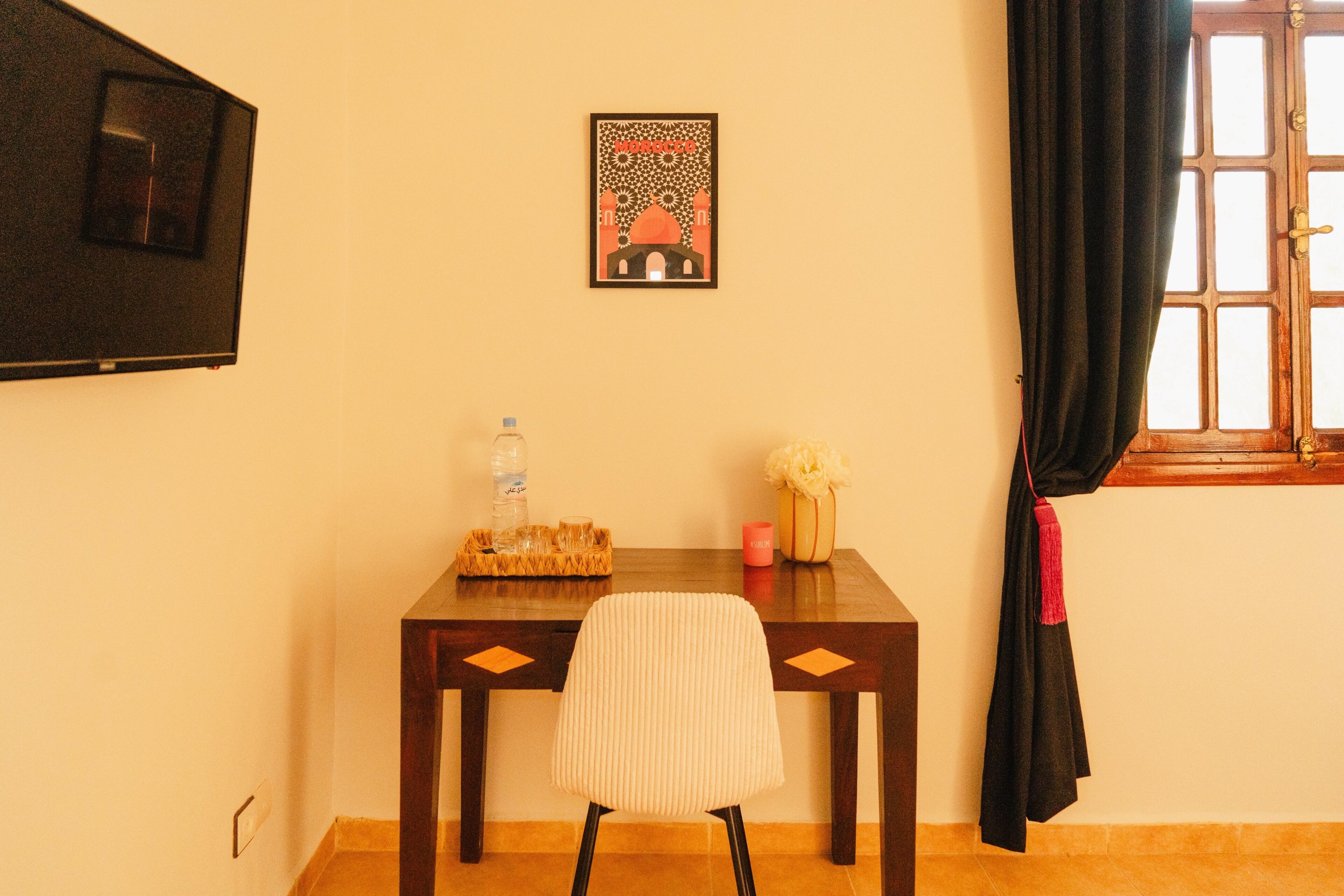 Ein Zimmer mit einem Holztisch, einem Stuhl, einem gerahmten Bild an der Wand, einem Fenster mit Vorhängen und einer Wasserflasche auf dem Tisch.