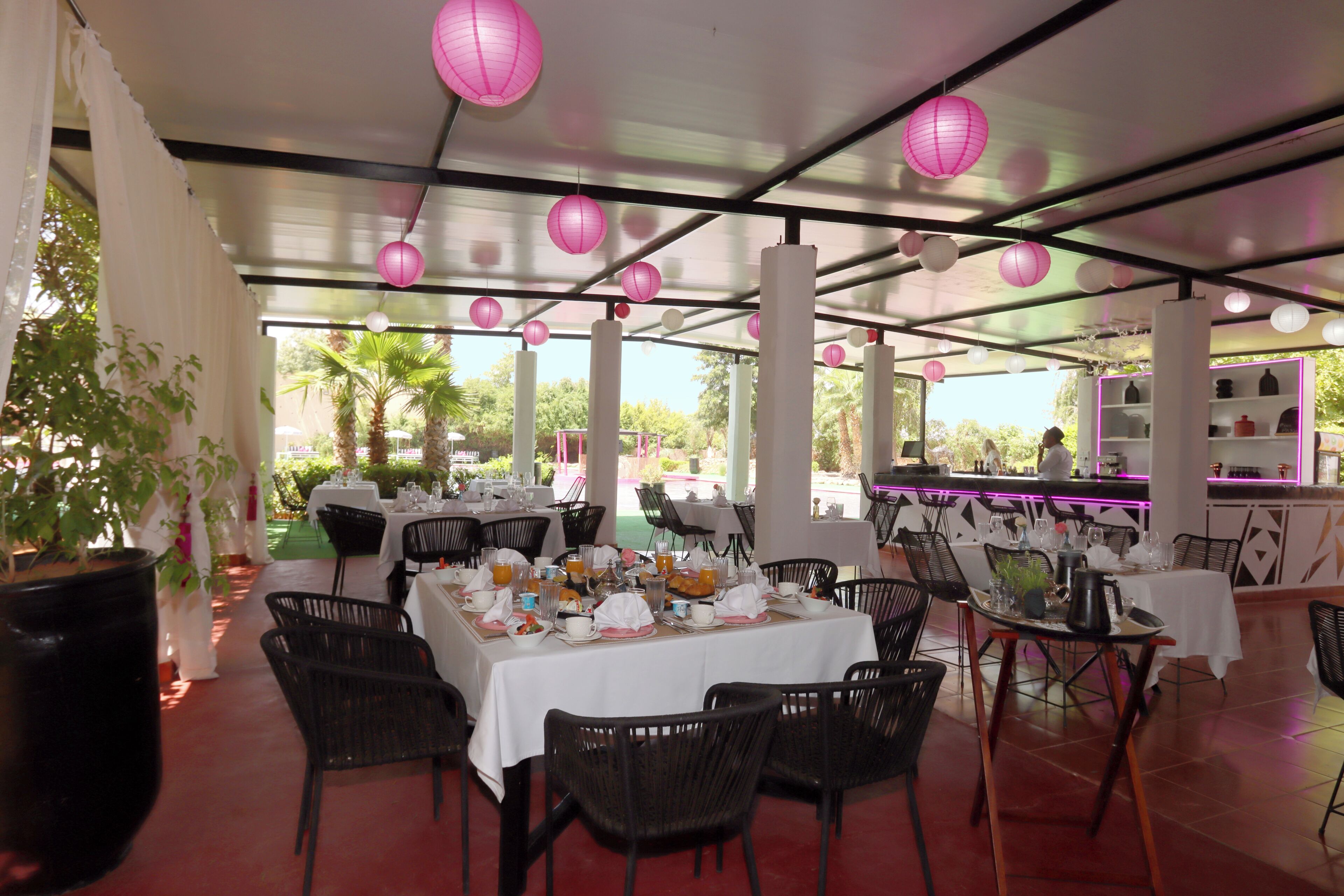 An outdoor dining area with tables set for breakfast, surrounded by chairs, and decorated with paper lanterns.