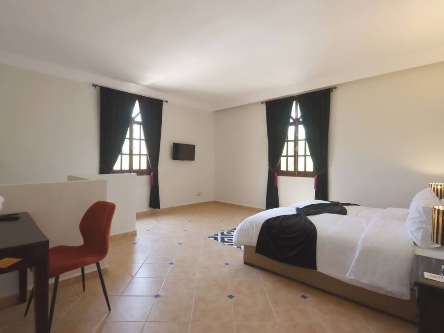 Une chambre d’hôtel avec un lit, un bureau, une chaise et deux fenêtres avec des rideaux.