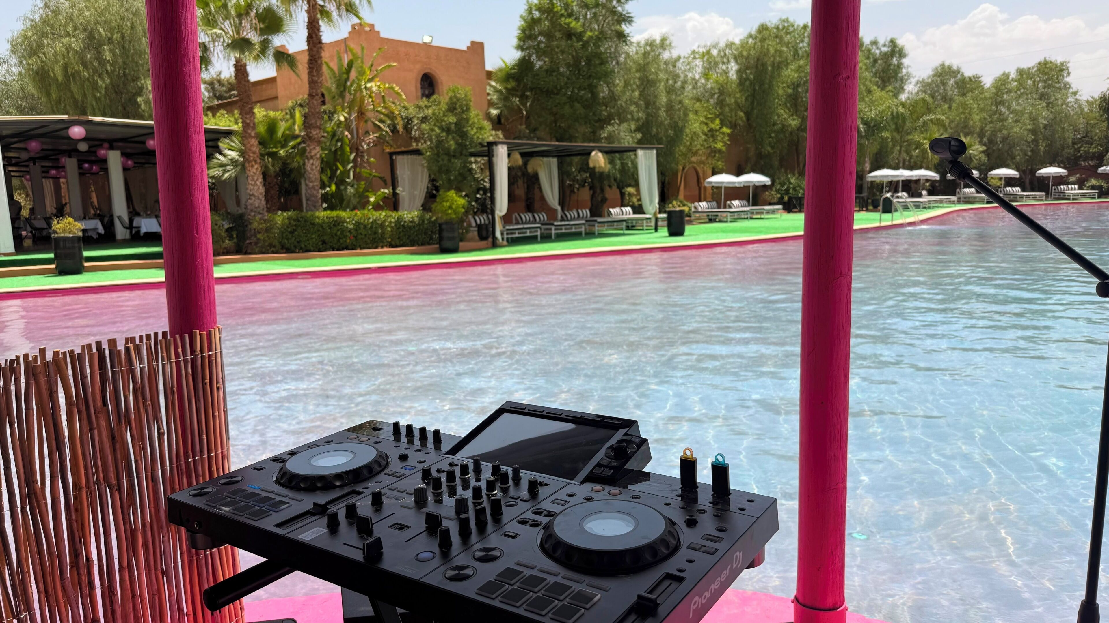 A DJ booth with a control panel, a pool with a pink border, and outdoor seating with umbrellas.
