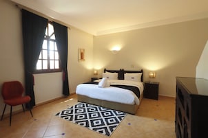 Una habitación de hotel con una cama, lámparas de noche, una silla roja, una alfombra estampada y un cuadro colgado en la pared.
