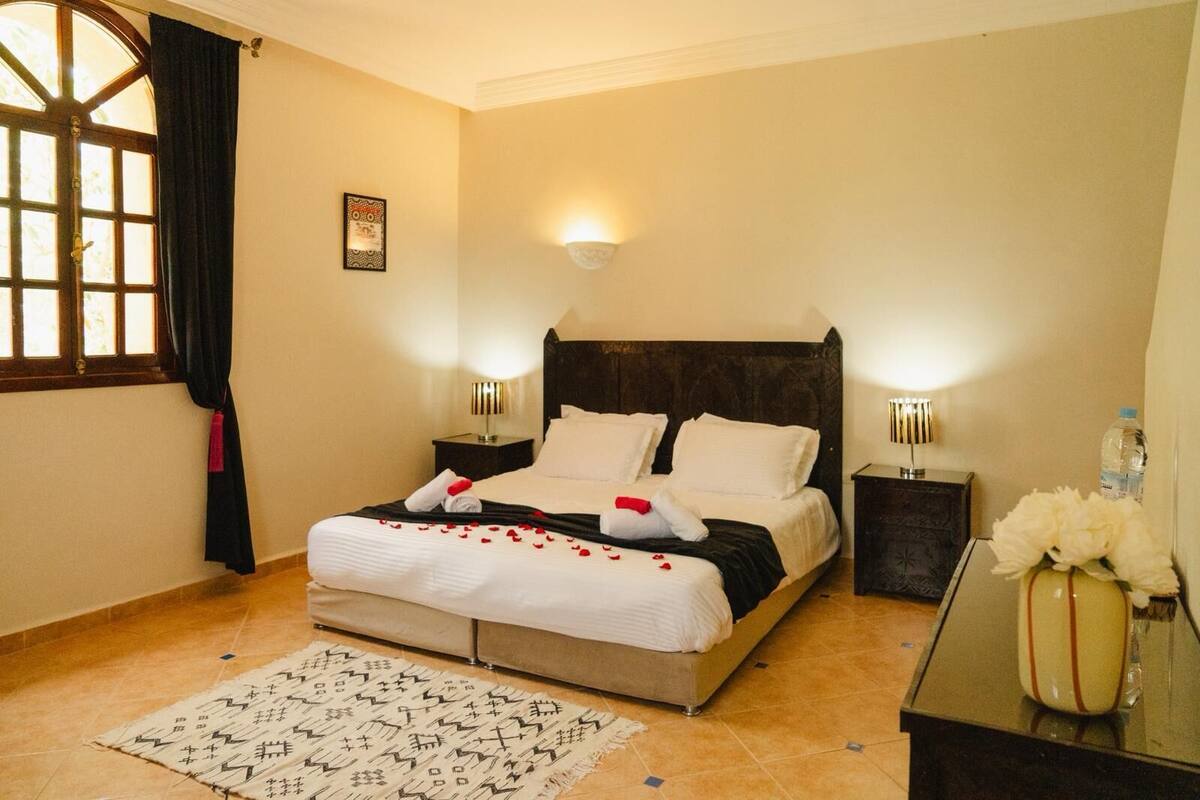 Une chambre d’hôtel comprenant un lit, des lampes de chevet, une table de chevet, une fenêtre avec des rideaux, un luminaire fixé au mur et un vase décoratif.