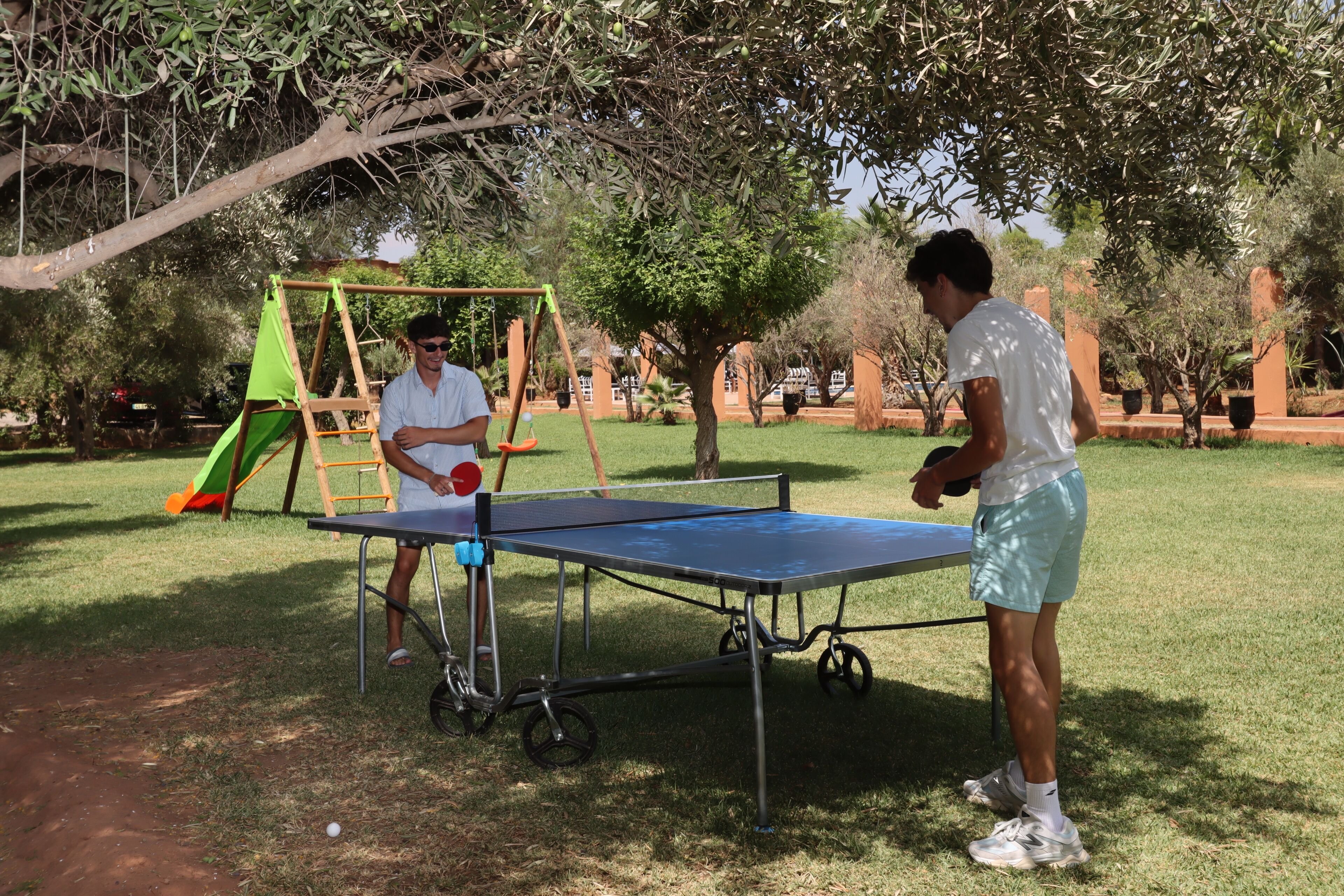An outdoor table tennis setup in a park with a swing set and trees in the background.