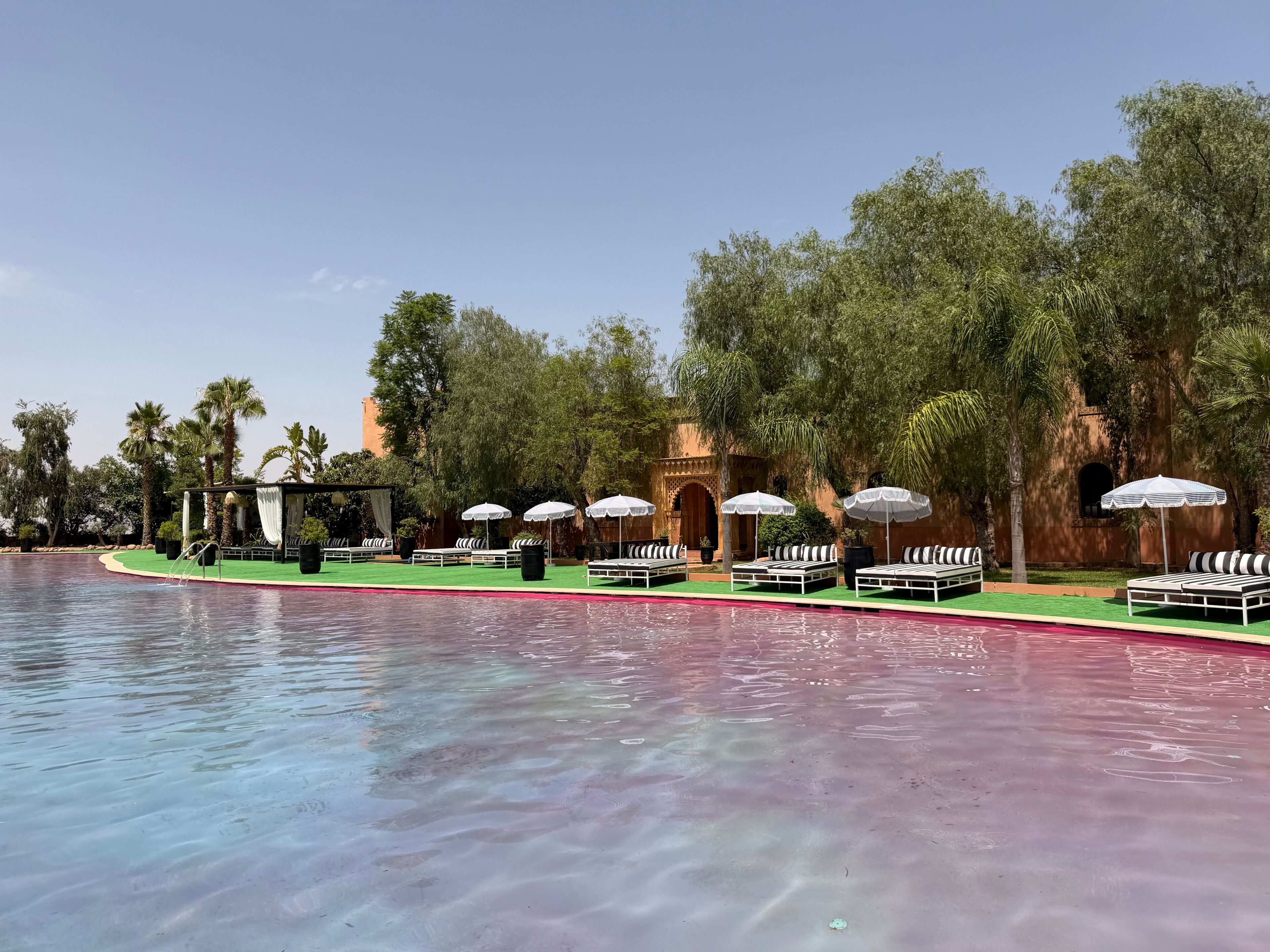 Zona junto a la piscina con sillones de descanso y sombrillas, rodeada de vegetación y un edificio con arcos.