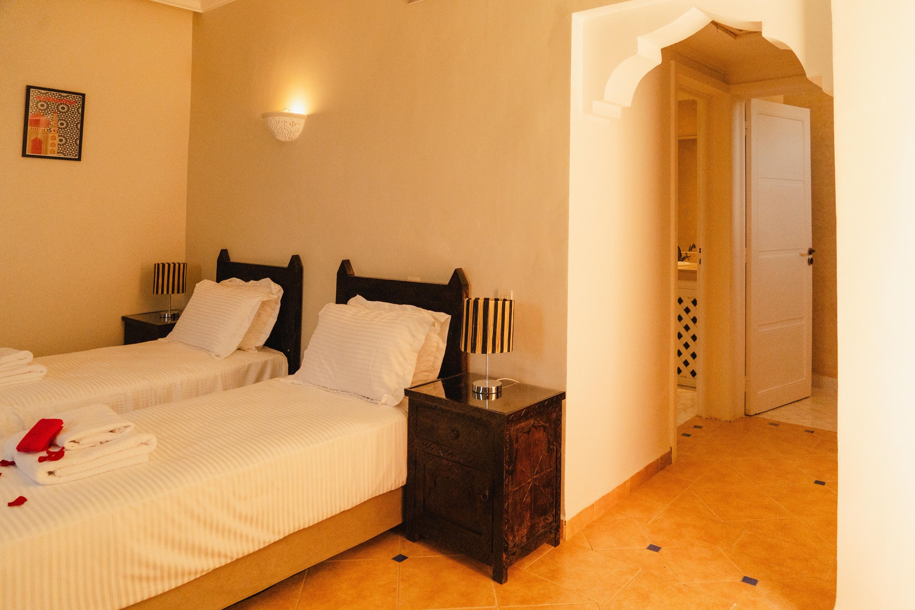 A hotel room with two beds, a nightstand, a wall-mounted lamp, and a door leading to another room.