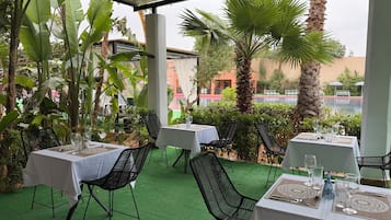An outdoor dining area with tables and chairs, surrounded by greenery and a view of a pool.