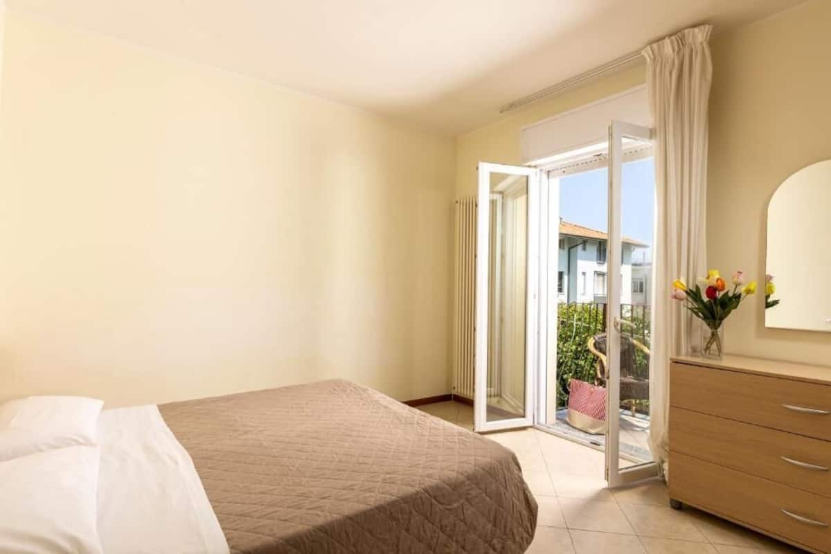 Une chambre d’hôtel comprenant un lit, une commode et une porte-fenêtre coulissante donnant sur un balcon aménagé avec du mobilier d’extérieur.