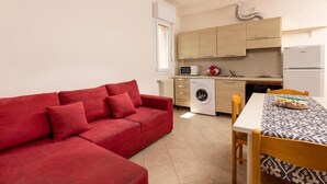 A compact living space with a red sofa, a dining table with a patterned tablecloth, a washing machine, and a kitchenette with wooden cabinets and a white refrigerator.
