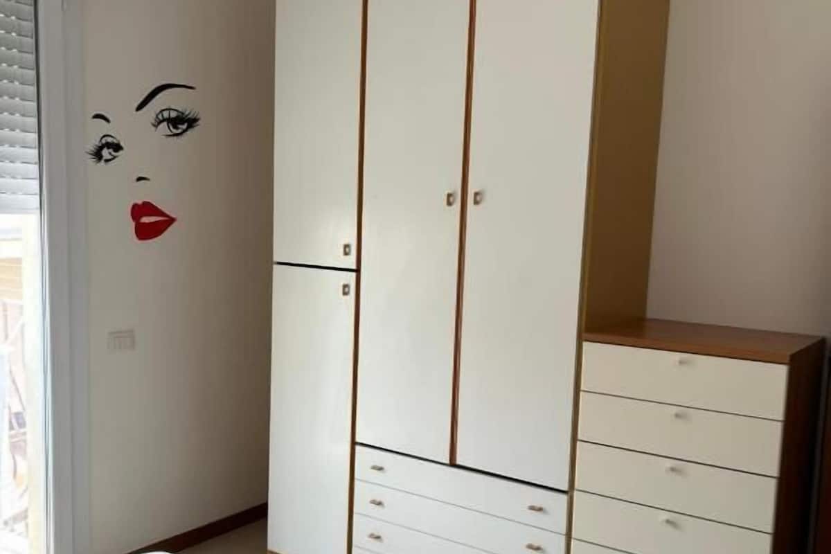Une chambre avec une grande armoire, un lit avec une literie à rayures et une fenêtre avec des stores.