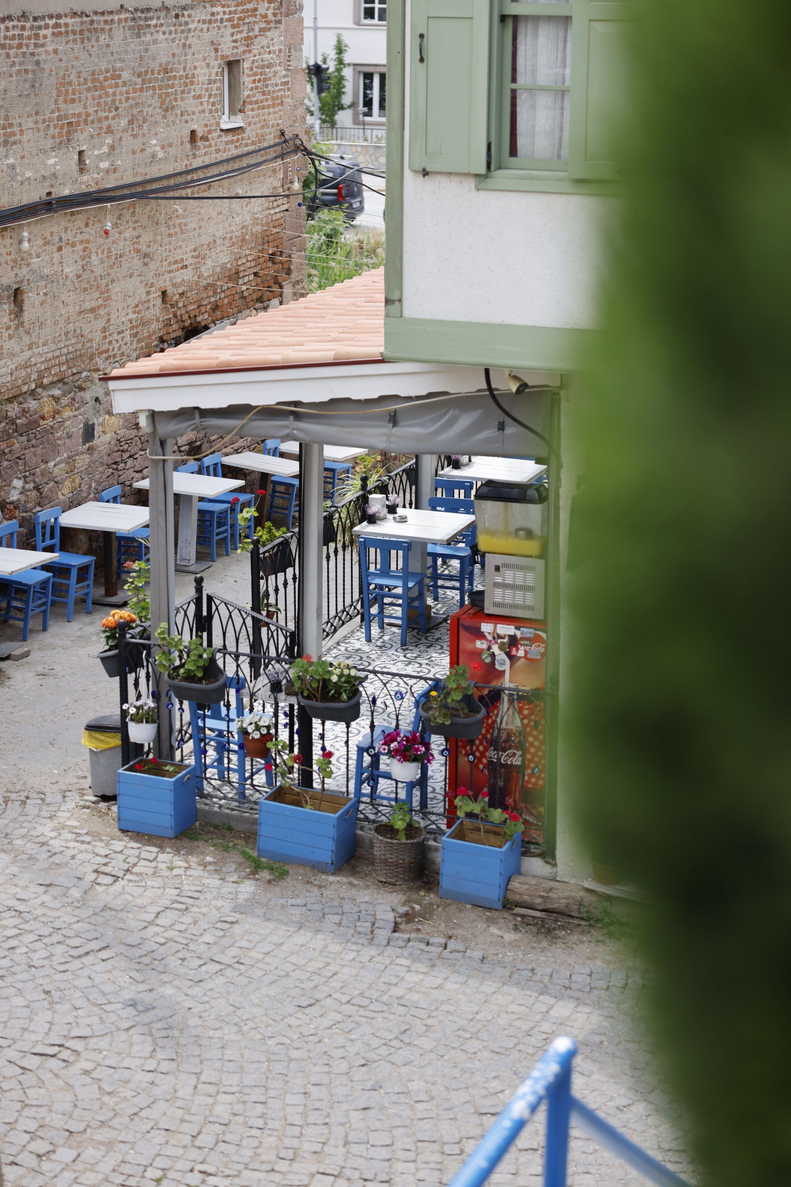 An outdoor café with tables and chairs, potted plants, and a brick wall in the background.