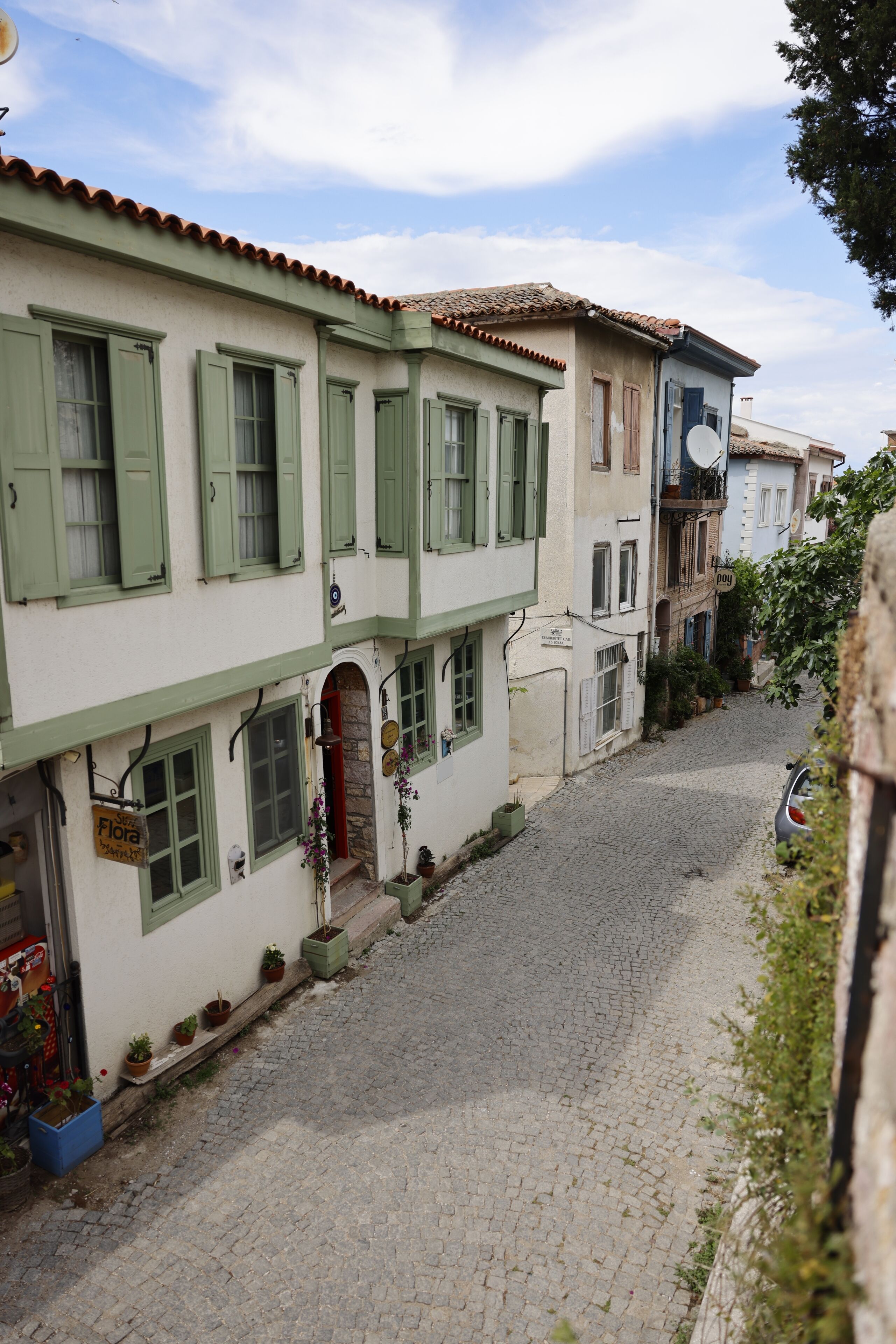 A narrow cobblestone street lined with buildings featuring green shutters and a sign for 'Flora.'