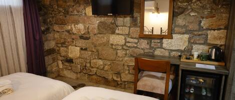A room with two beds, a television mounted on a stone wall, a mirror, a wooden chair, and a minibar.