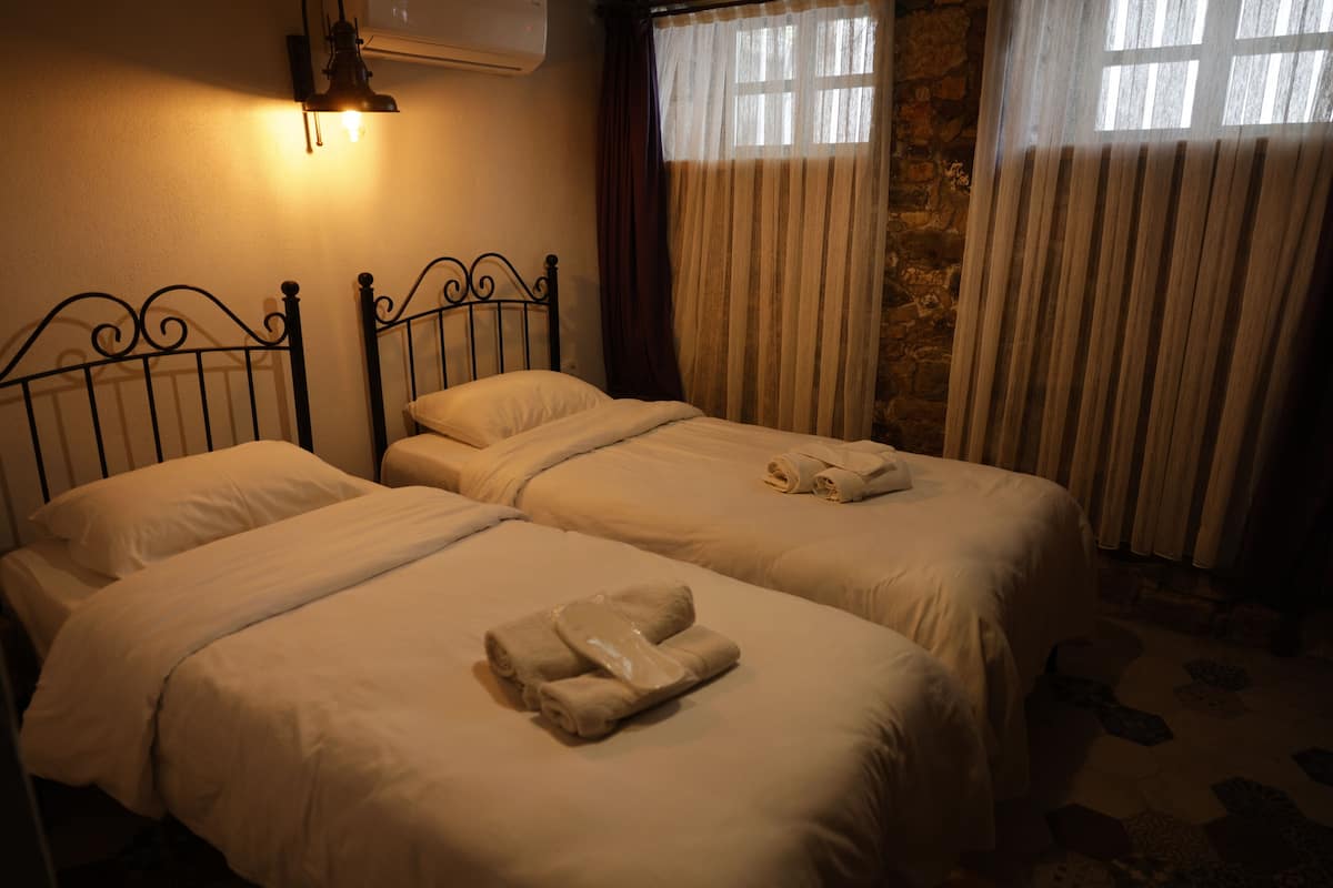 Dos camas con sábanas y toallas blancas, un aire acondicionado de pared y una ventana con cortinas.