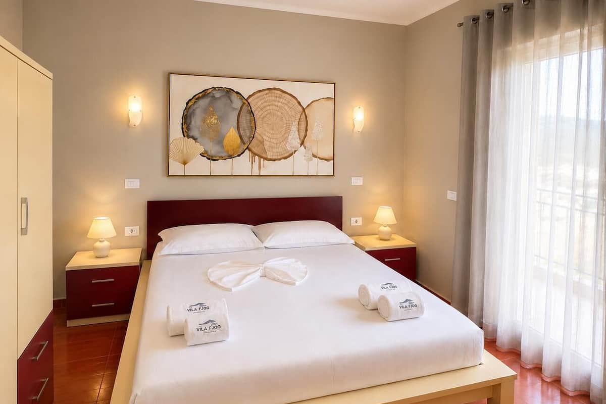 Habitación de hotel con cama, dos mesitas de noche con lámparas, un cuadro en la pared y una ventana con cortinas.