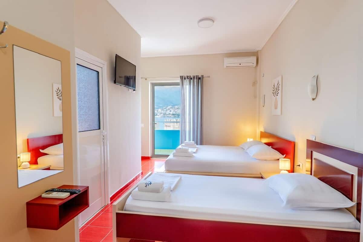 Habitación de hotel con dos camas, televisión, balcón con vistas y una pared de acento roja.