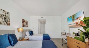 Ein Hotelzimmer mit zwei Betten, einem Schreibtisch, einem Sessel, einem Fernseher und einem Bild mit Strandmotiv.