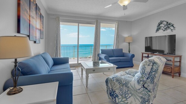 A living room with a sofa, armchair, coffee table, TV, and a balcony with a view of the ocean.