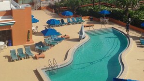 A swimming pool with lounge chairs and umbrellas, a beach view, and a building with balconies.