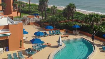 A swimming pool with lounge chairs and umbrellas, a beach view, and a building with balconies.