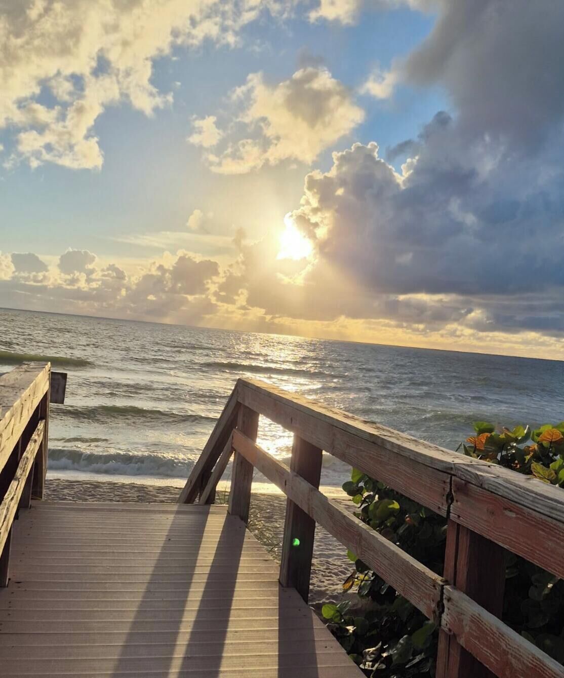 A wooden pier leading to a beach with ocean waves and a partly cloudy sky.