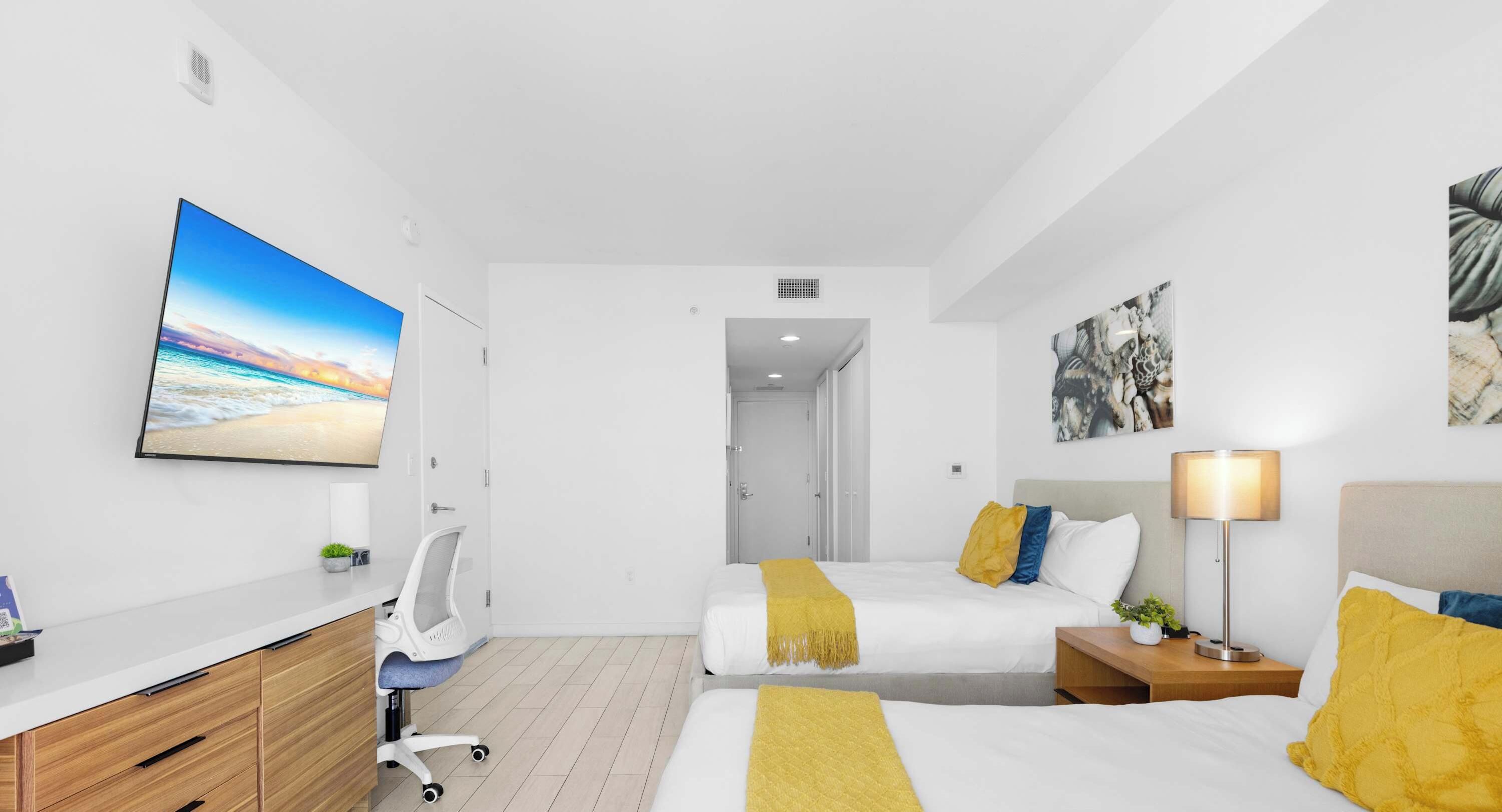 A hotel room with a bed, bedside table, lamp, desk, chair, and artwork on the walls.
