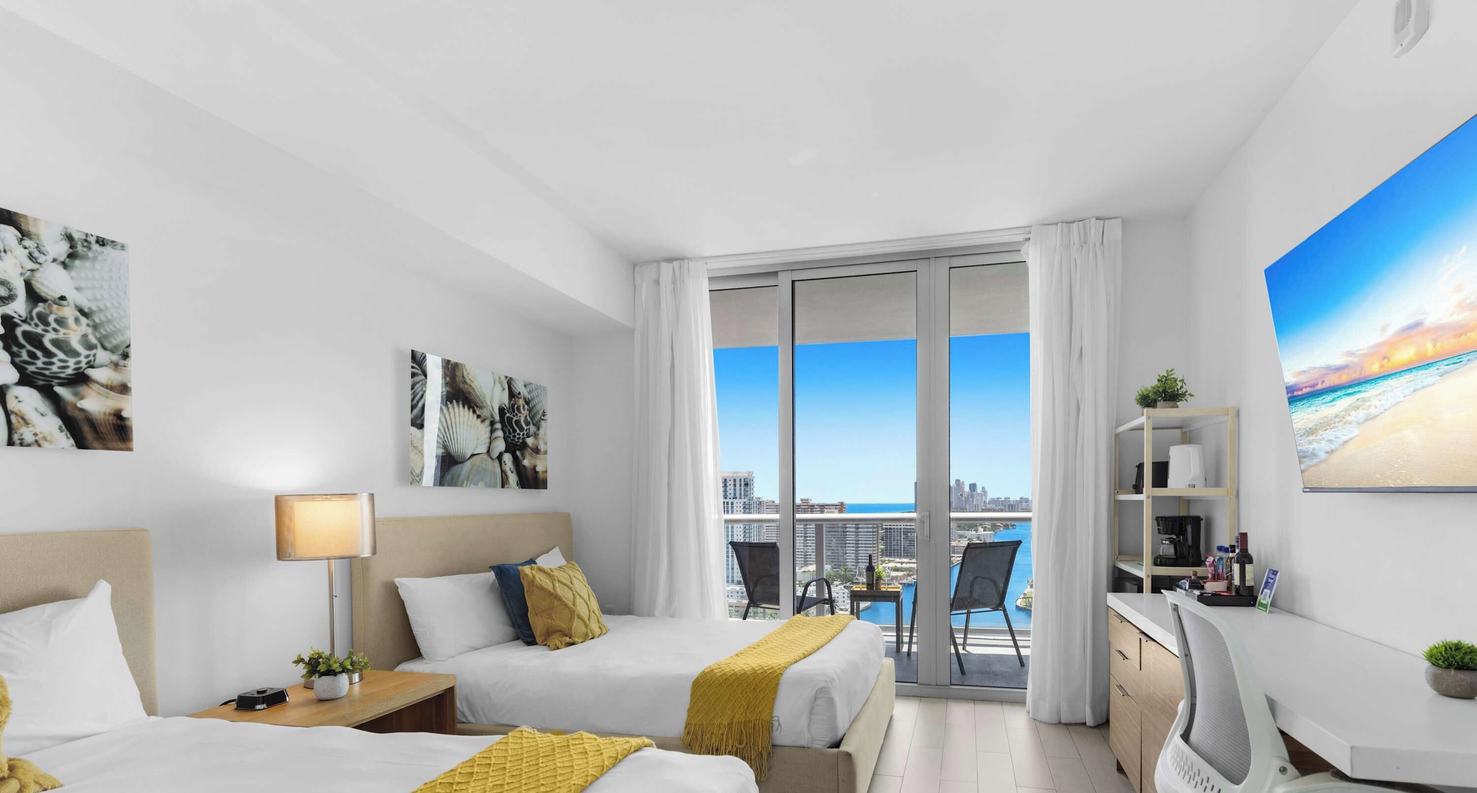 Habitación de hotel con dos camas, un escritorio, una silla, balcón con vista a la ciudad y obras de arte en las paredes.