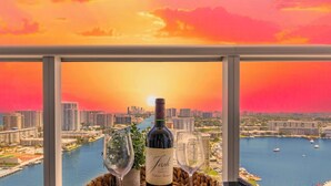 A table with a bottle of wine and two glasses, set on a balcony overlooking a cityscape and a body of water during sunset. - Intracoastal View Apt | Balcony & Beach Shuttle (Hallandale Beach)
