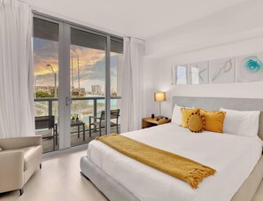 A hotel room with a bed, two bedside lamps, a nightstand, a view of the city through large windows, and a balcony with a chair. - Marina Views  2BR  Beachwalk Resort (Hallandale Beach)