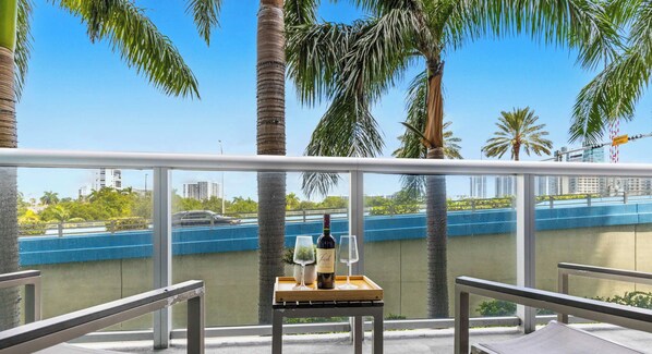 A balcony with a table set for two, overlooking a blue waterfront and palm trees. - Marina Views  2BR  Beachwalk Resort (Hallandale Beach)