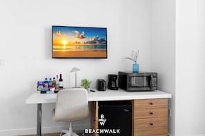 Ein modernes Zimmer mit Schreibtisch, Stuhl, Mikrowelle, Kaffeemaschine, einer Pflanze und einem gerahmten Bild vom Strand bei Sonnenuntergang.