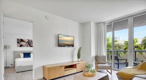 A modern living room with a large window, a flat-screen TV, a sofa, a chair, and a wooden cabinet. - Cozy 2BR Apartment · Intracoastal · Beachwalk (Hallandale Beach)
