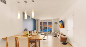 Ein modernes Wohnzimmer mit Essbereich, großen Fenstern und Blick auf die Stadt sowie einem an der Wand befestigten Flachbildfernseher.