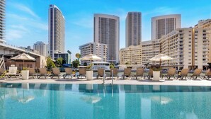 Area piscina di un hotel con lettini e ombrelloni, circondata da edifici di grandi dimensioni.