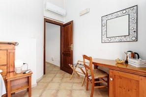 Una stanza con una scrivania in legno, una sedia e un armadietto, una porta aperta che dà su un'altra stanza e uno specchio fissato alla parete.