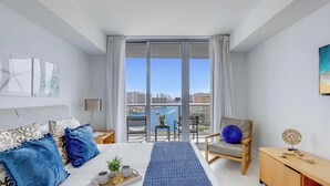 Quarto moderno com cama grande, uma cadeira, mesa de cabeceira e vista para a cidade através de uma varanda.