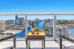 A balcony with a clear view of a cityscape, a body of water, and a bottle of wine on a tray. - Intracoastal Panorama · 2BR Beachwalk Resort (Hallandale Beach)