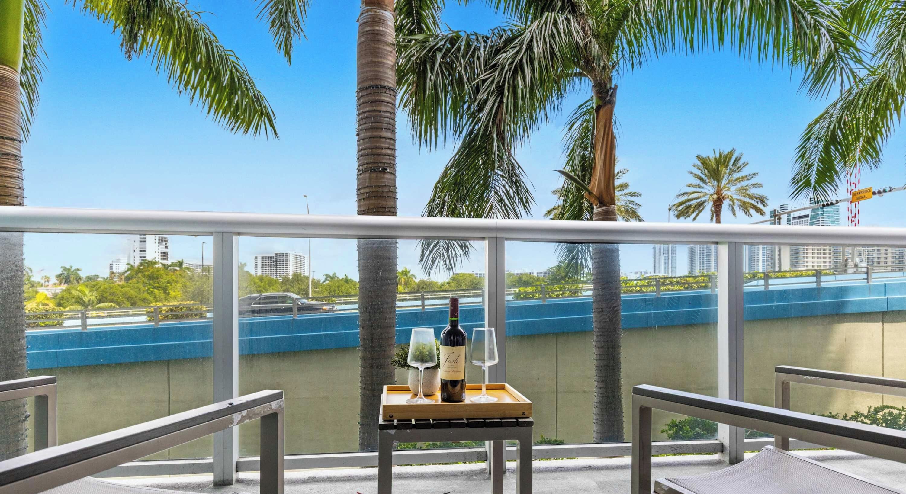 Varanda com mesa posta para dois, com vista para a frente marítima azul e palmeiras.