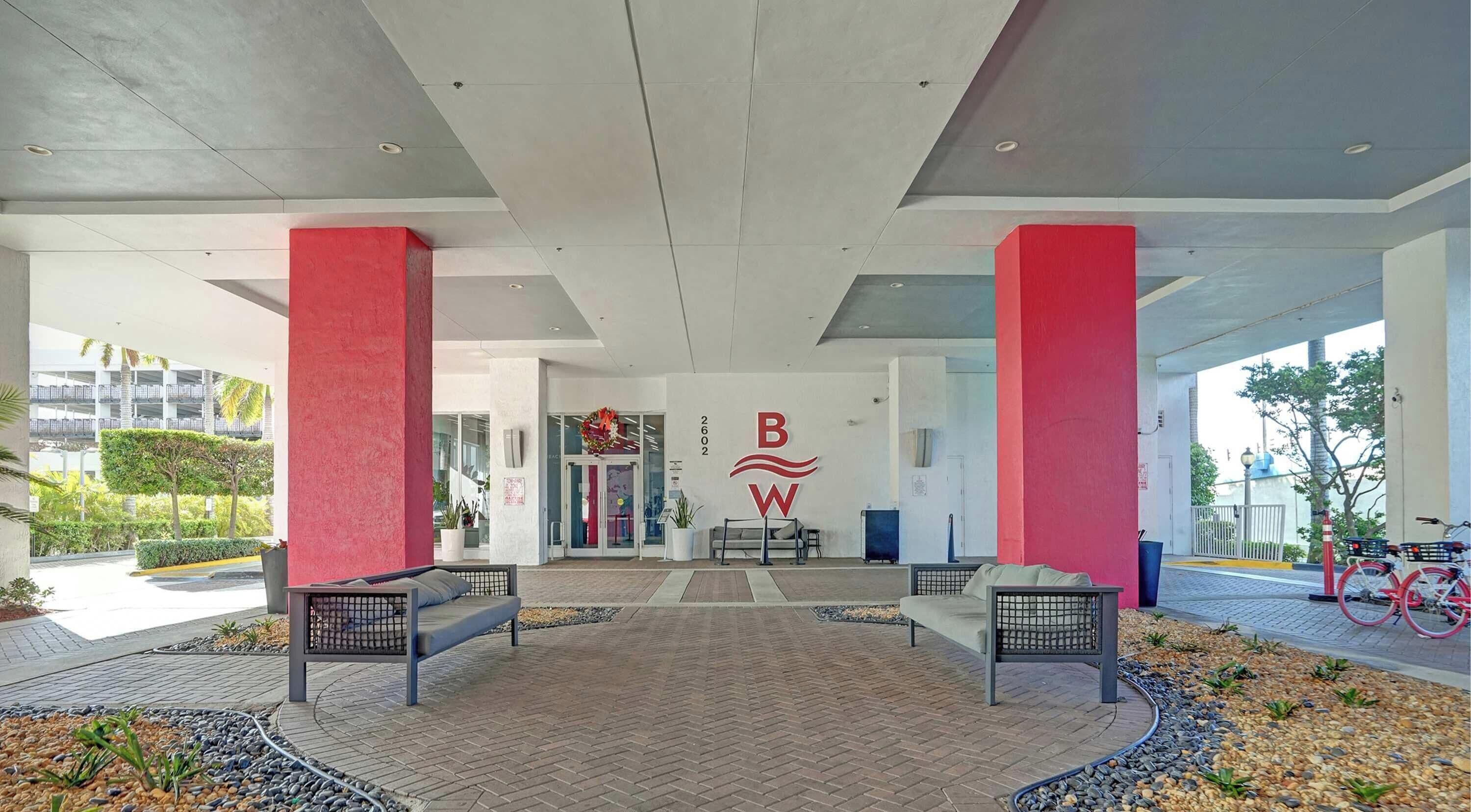 A modern building entrance with a red and white logo, two benches, and a paved area with plants.