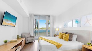 A hotel room with a bed, a desk, a chair, a nightstand, and a view of the outdoors. - Spacious BW - 1BR APT - Resort‑Style experience (Hallandale Beach)