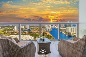 Ein Balkon mit Korbmöbeln, einer Flasche und Gläsern, mit Blick auf eine Stadtlandschaft mit Fluss und Sonnenuntergang.